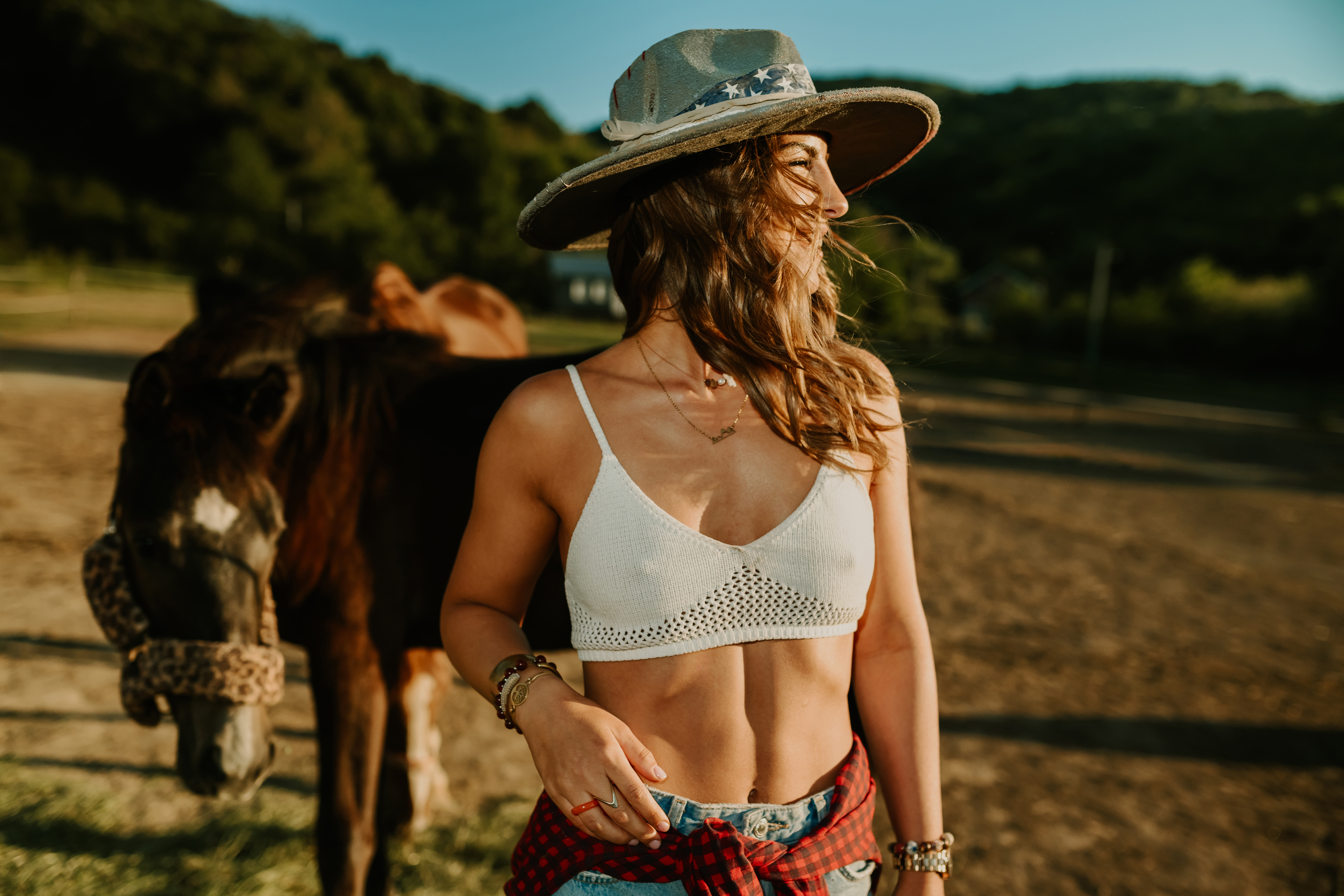 Portrait of a young handsome rancher girl in western styled clothes and cowboy hat standing at horse pasture with horses grazing in a blurry background. Smiling cowgirl is posing at horse ranch.
